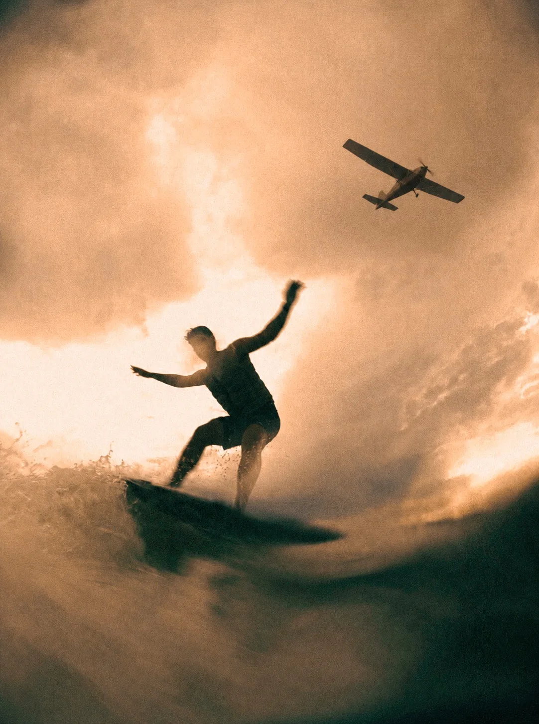 Surfer riding a wave beneath a small airplane at sunset